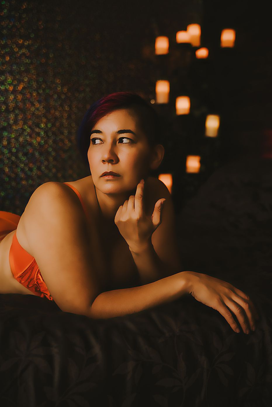 Moody boudoir portrait in warm lighting with floating candles creating romantic ambiance in dark setting.