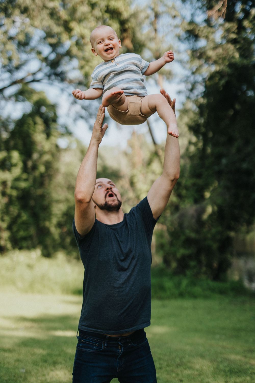 A person in a black t-shirt playfully tosses a small child in the air at a sunny outdoor park.