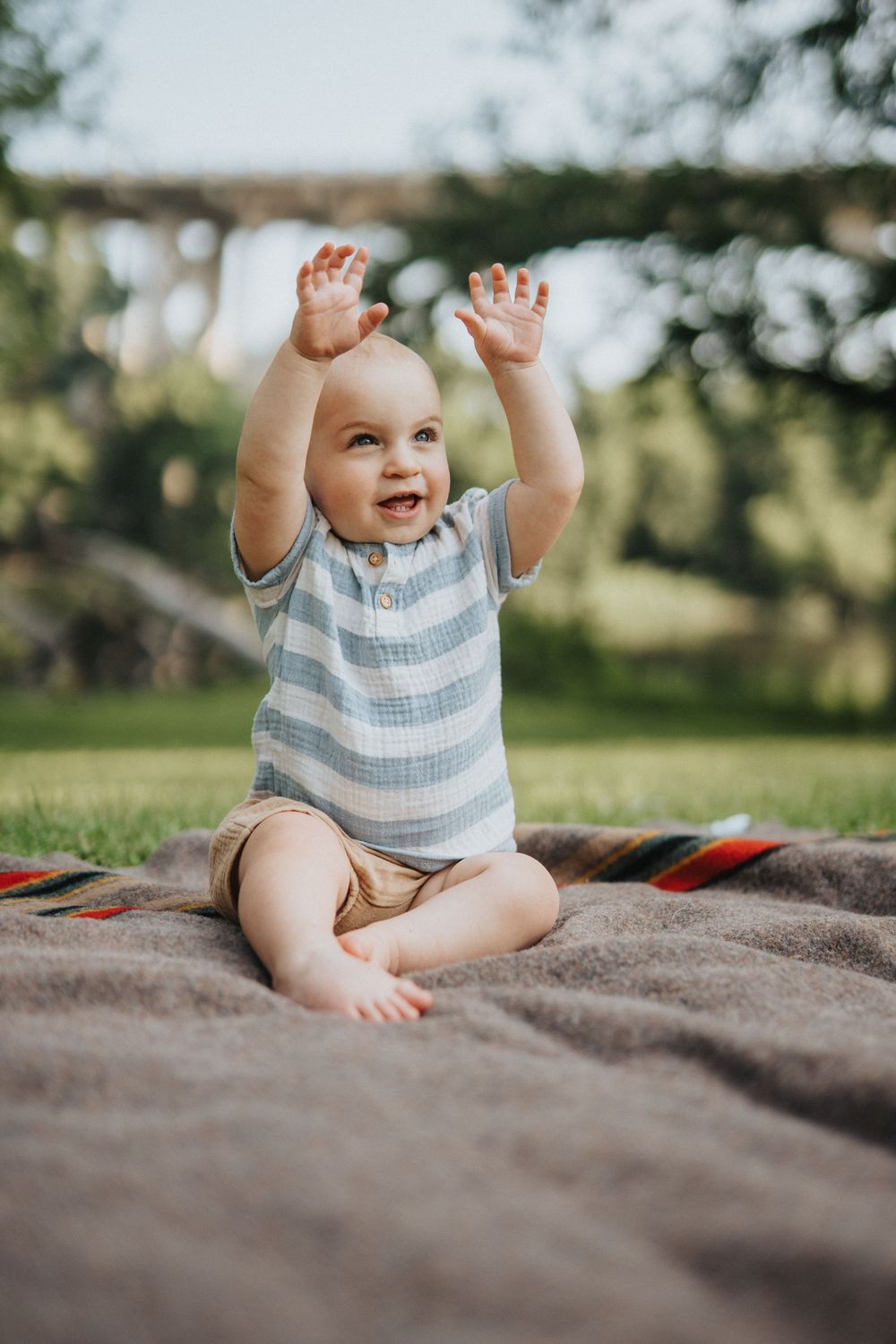 Baby in striped onesie sits on blanket outdoors raising hands joyfully in sunlit garden setting.