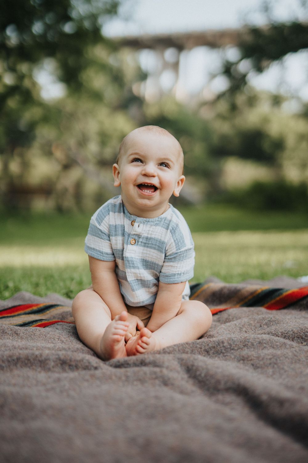 Baby in striped onesie sits and laughs on a blanket outdoors during a sunny photo session.