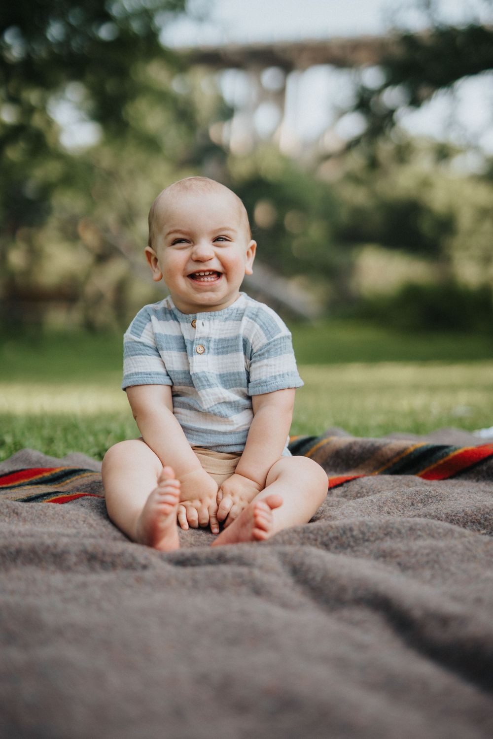 Baby in striped shirt sits on a blanket outdoors with a joyful expression.