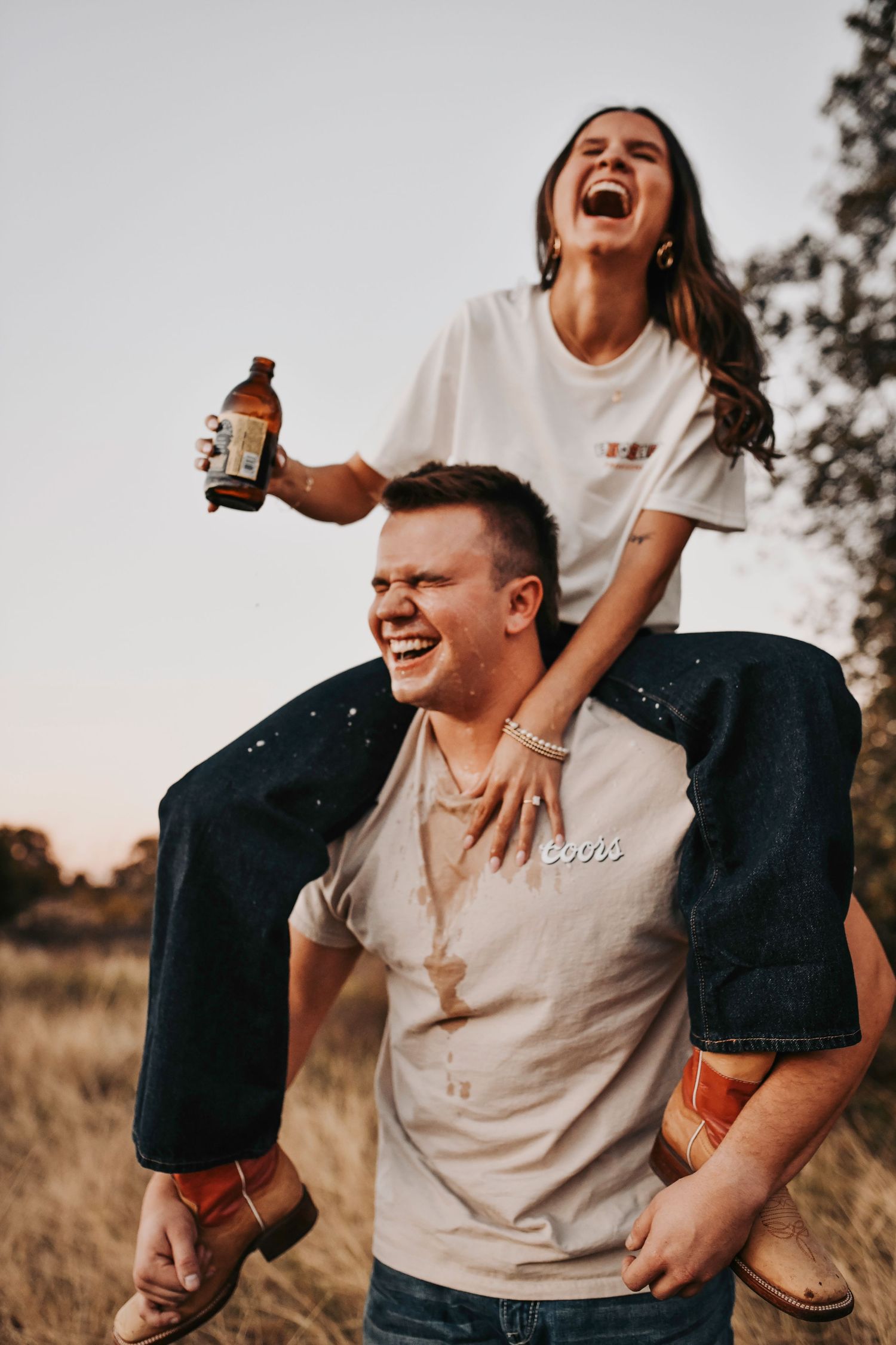 Fun engagement photos of a firefighter lifting his fiancée on his shoulders while they laugh and hold beers
