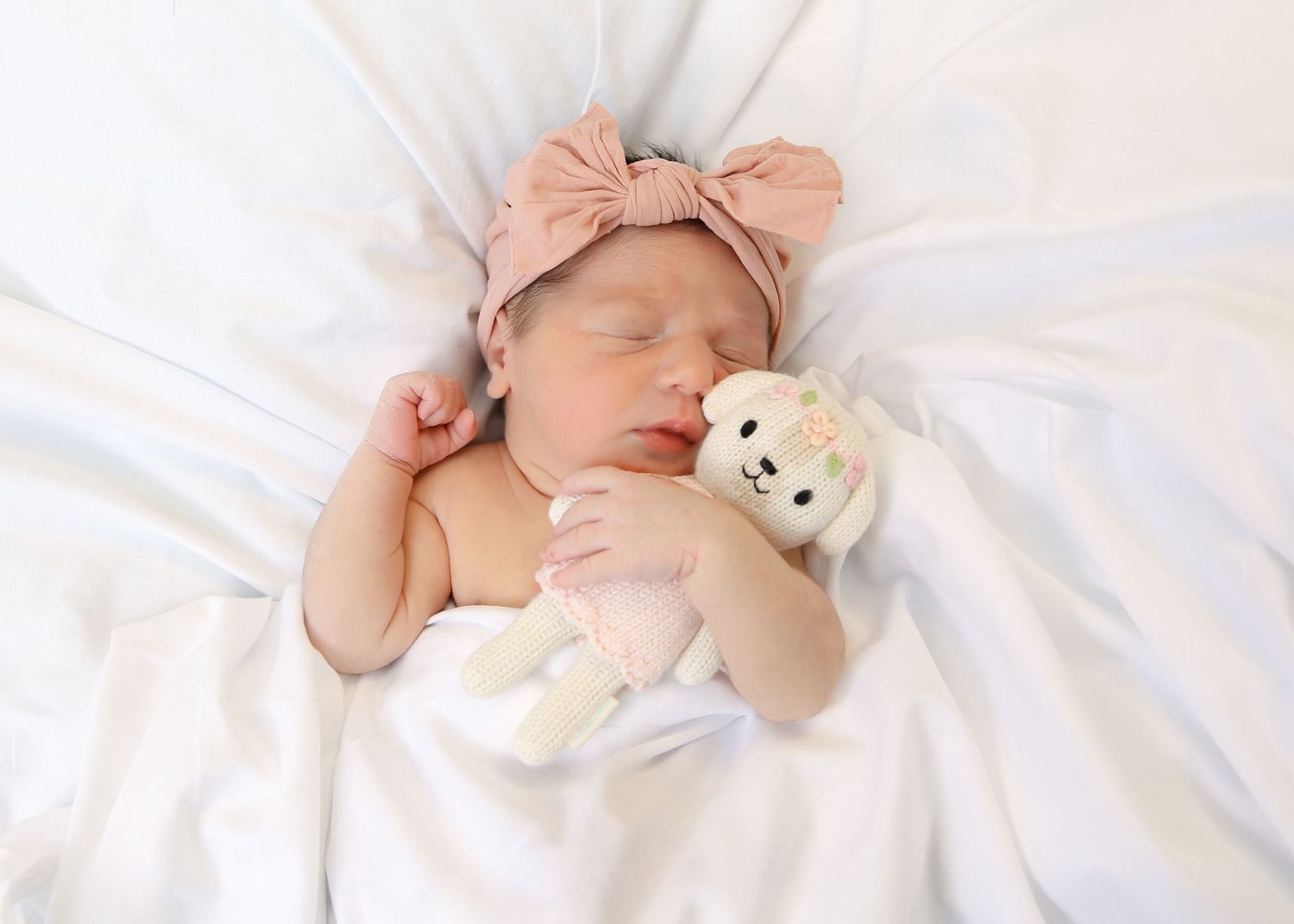 A newborn baby sleeps peacefully in white bedding while holding a small teddy bear and wearing a pink bow.