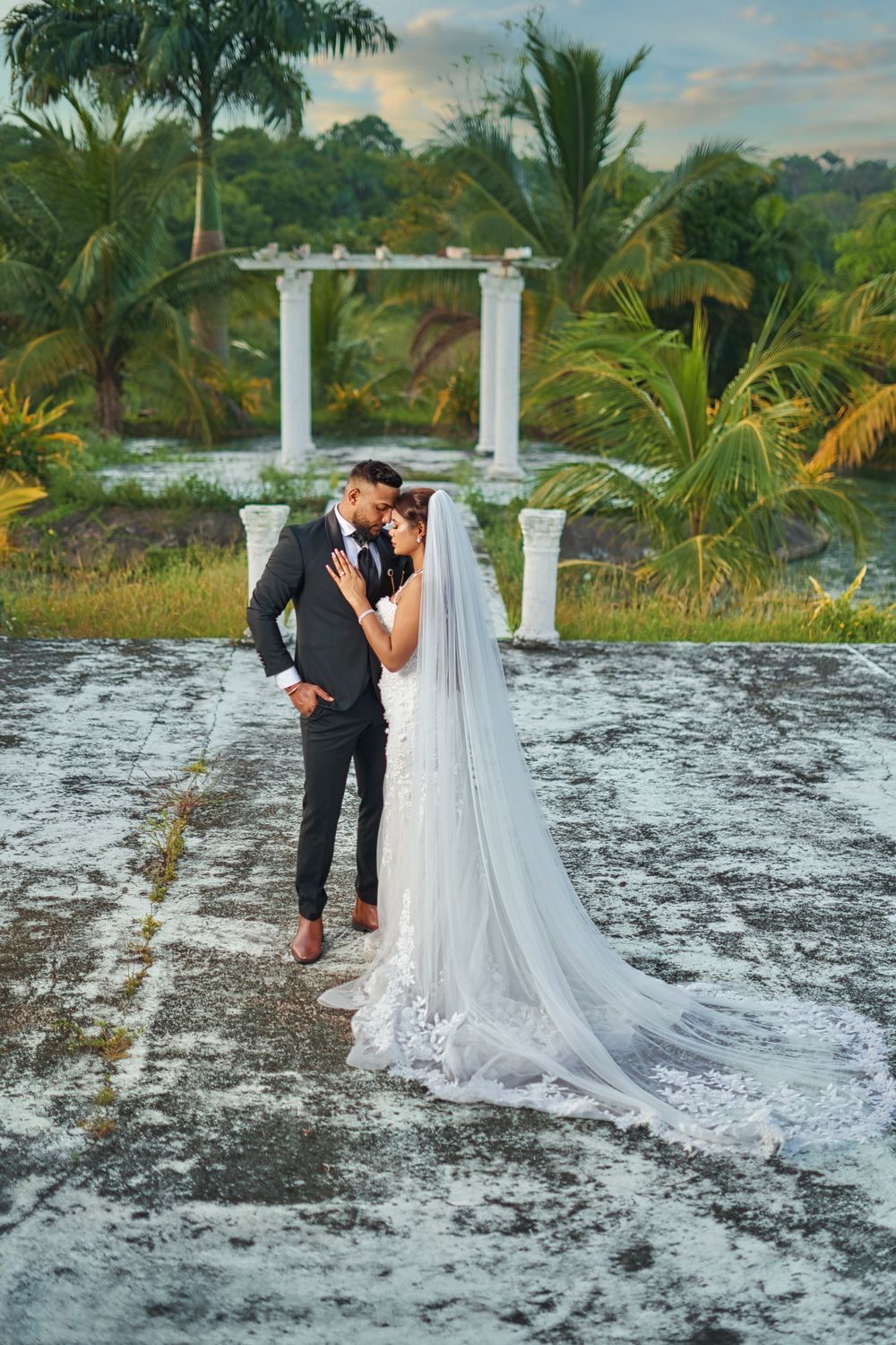 A couple in formal wedding attire poses together on a snowy beach with palm trees and tropical scenery in the background.