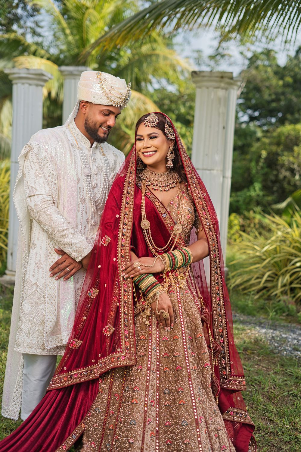 A wedding couple in ornate Indian attire poses together against a backdrop of palm trees and white pillars.