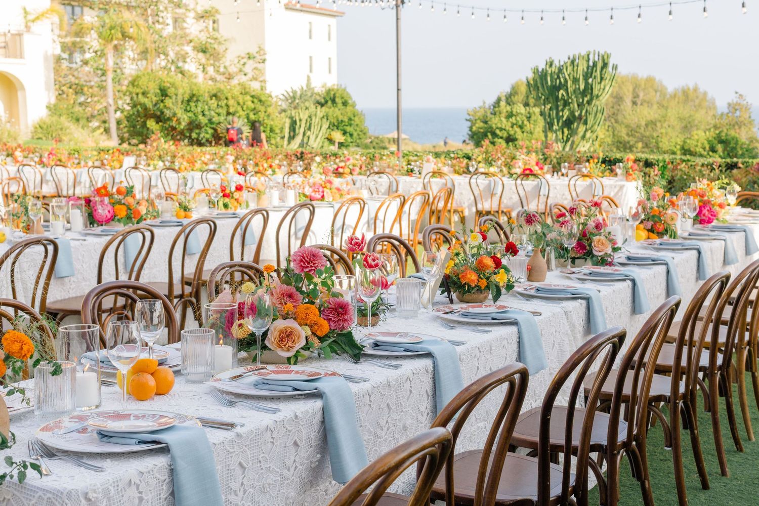 Elegant outdoor wedding reception setup with rustic wooden chairs and floral centerpieces on white tablecloths at sunset.