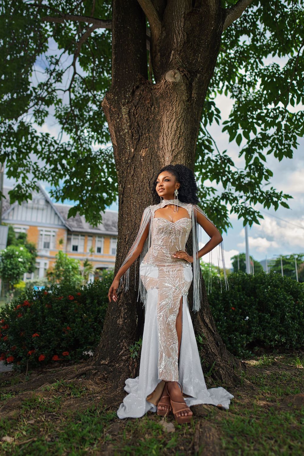 Elegant white lace wedding dress with flowing cape photographed against a large tree in an outdoor garden setting.