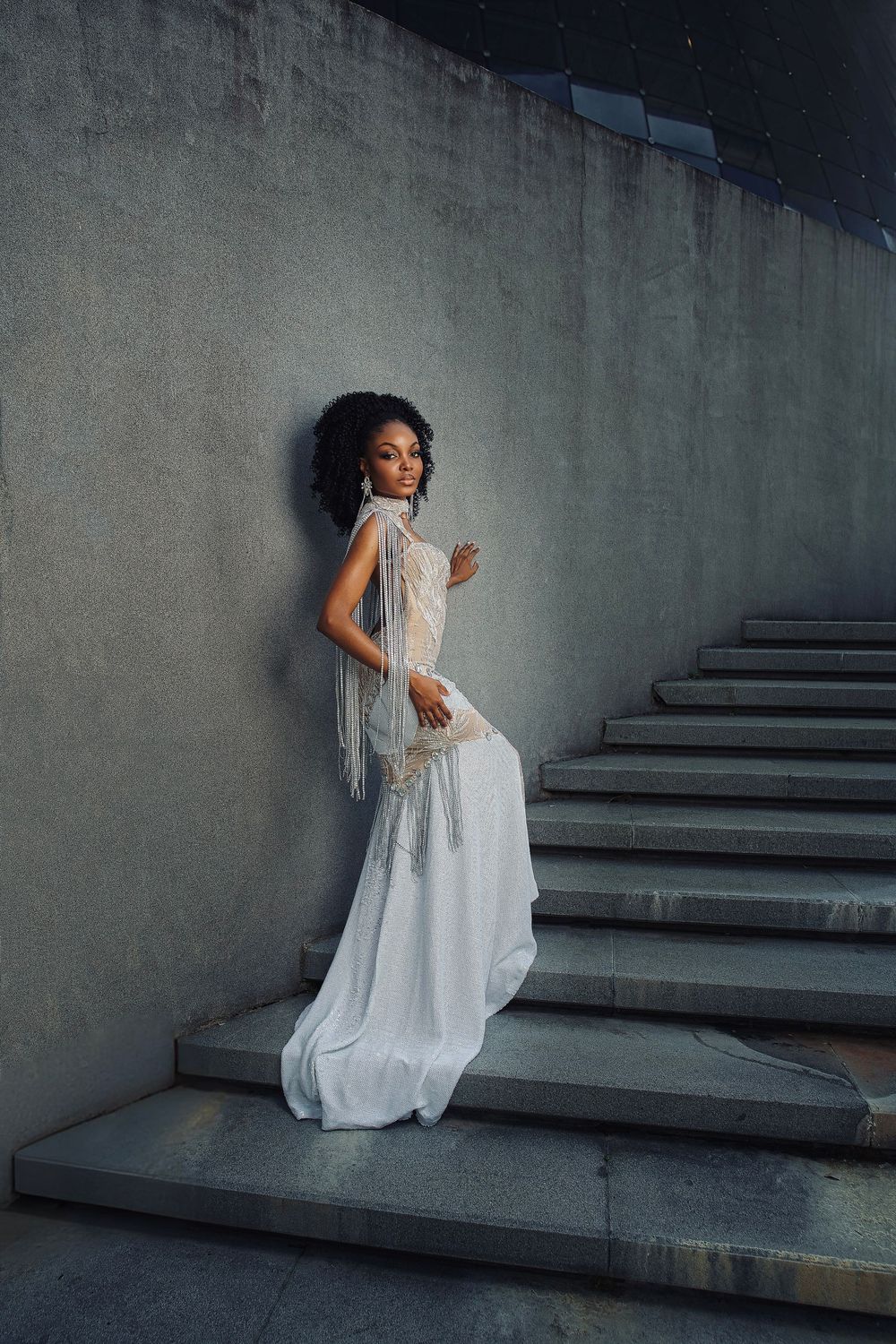 A figure in a flowing white dress poses gracefully on concrete steps against a moody gray wall.