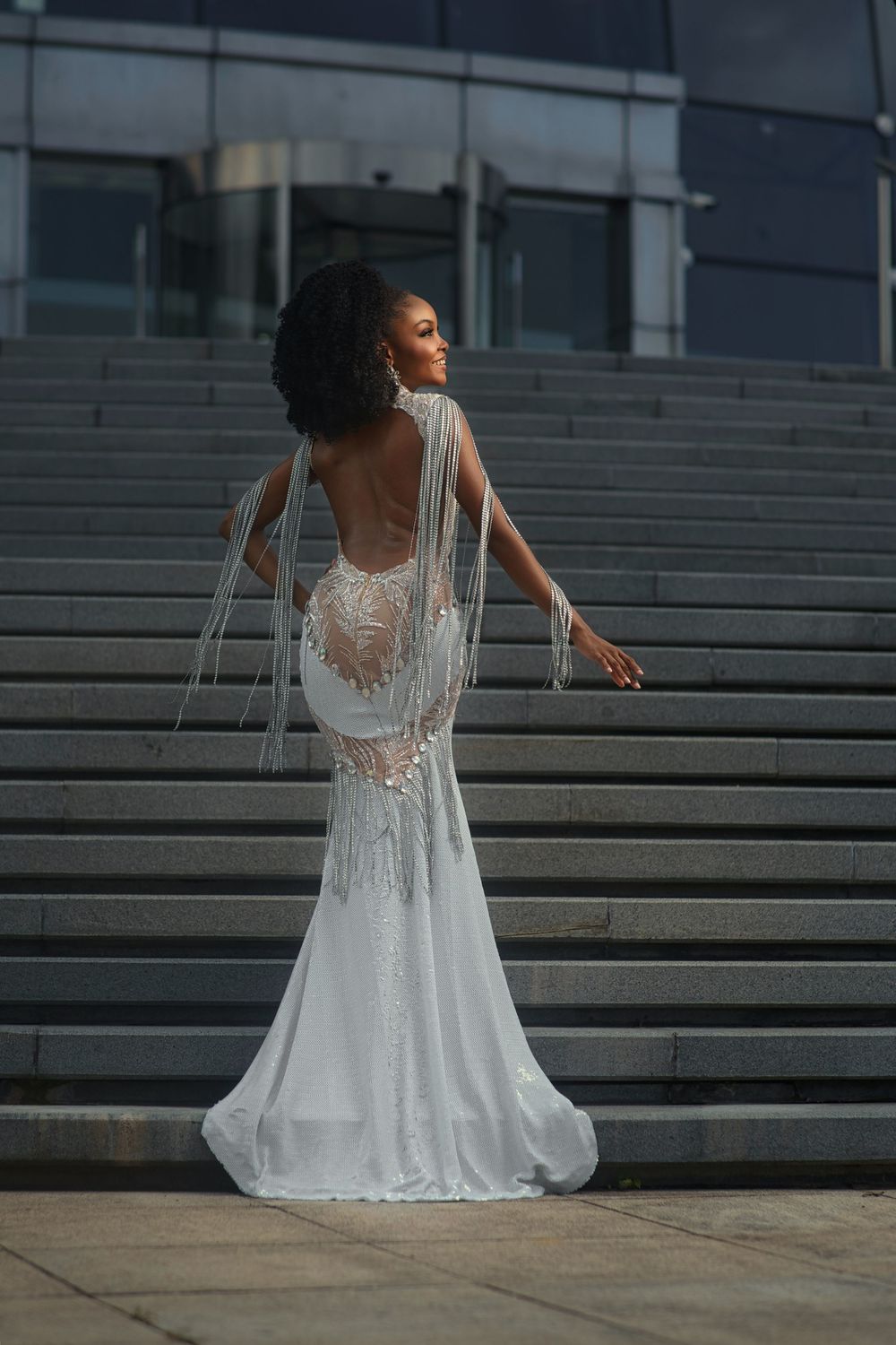 A bride in an elegant white lace wedding gown with flowing cape details poses gracefully on modern concrete steps at dusk.