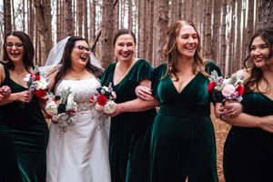 A wedding party in emerald green velvet dresses and white gown stands laughing together in a pine forest. Oak Openings Metropark.