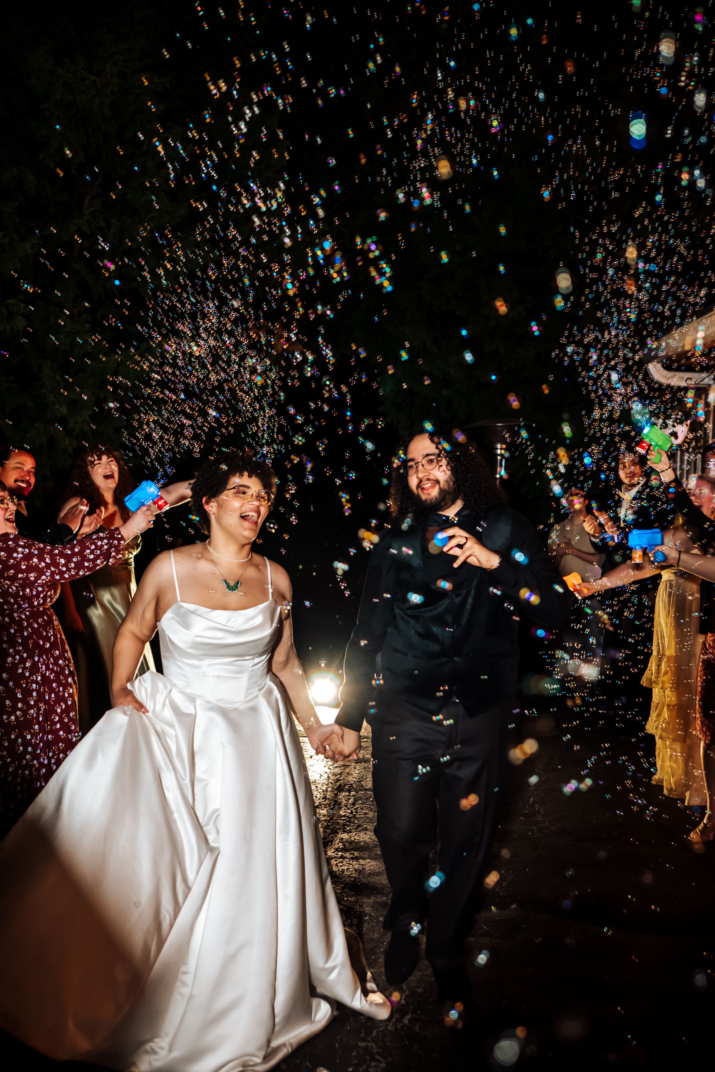 Joyful newlyweds walk through a colorful confetti shower at night, surrounded by cheering guests at their wedding celebration.