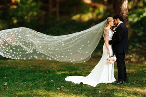 Newlywed couple embraces in a sunlit garden as the bride's long veil flows dramatically in the breeze at Nazareth Hall.