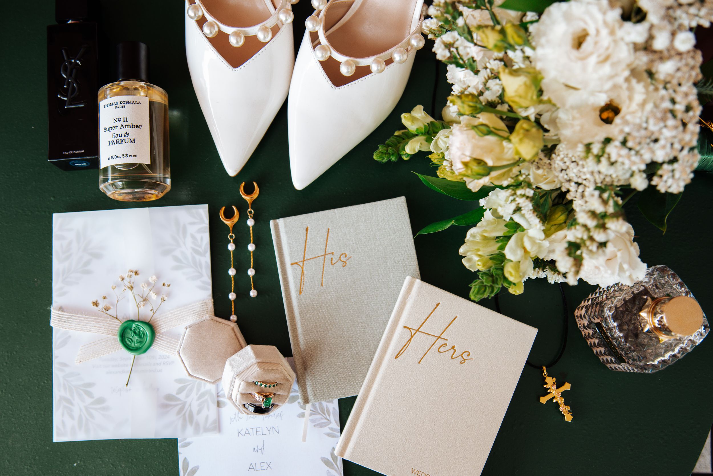 Wedding details including white shoes, floral arrangements, and elegant stationery laid out on a dark green surface.