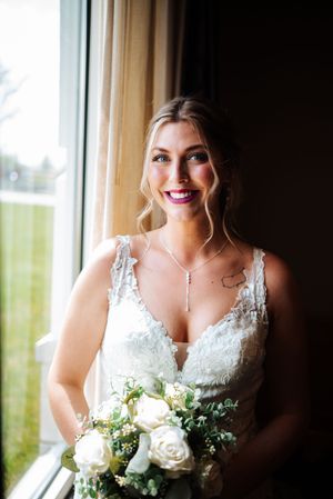 A bride in a white lace wedding dress holds a bouquet of white roses and greenery while standing by a window.