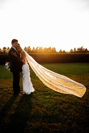 A couple embraces in a golden sunset field with a long flowing cathedral veil catching the warm evening light.