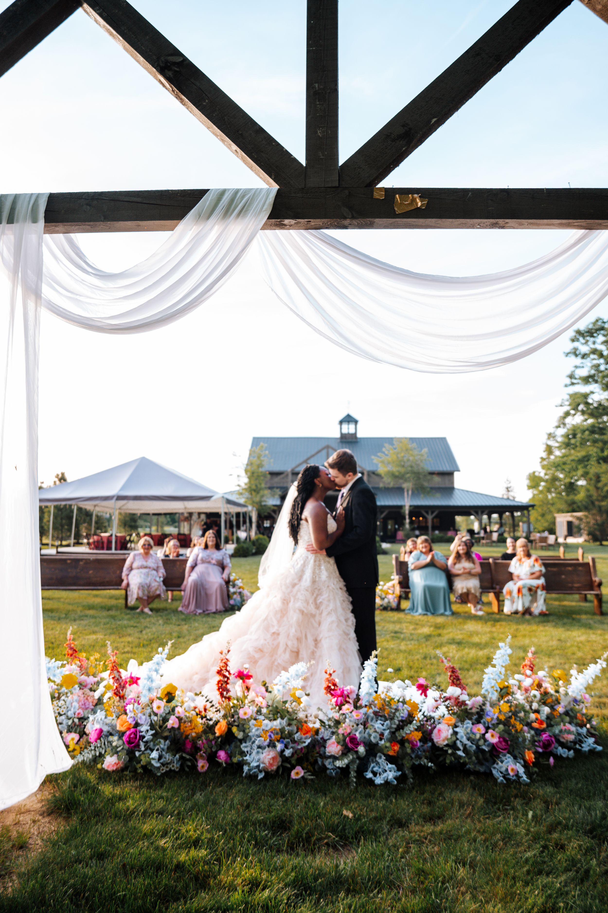 Bride and groom share first kiss under floral arch with colorful blooms at outdoor barn wedding ceremony.