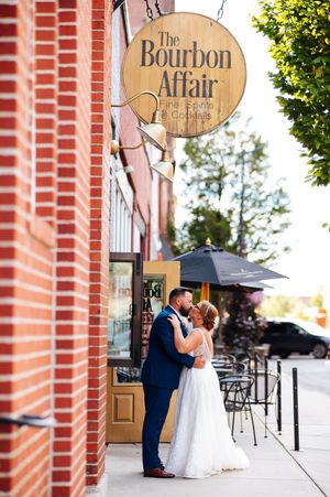 A romantic wedding couple shares a tender moment in front of a rustic brick building with vintage signage.