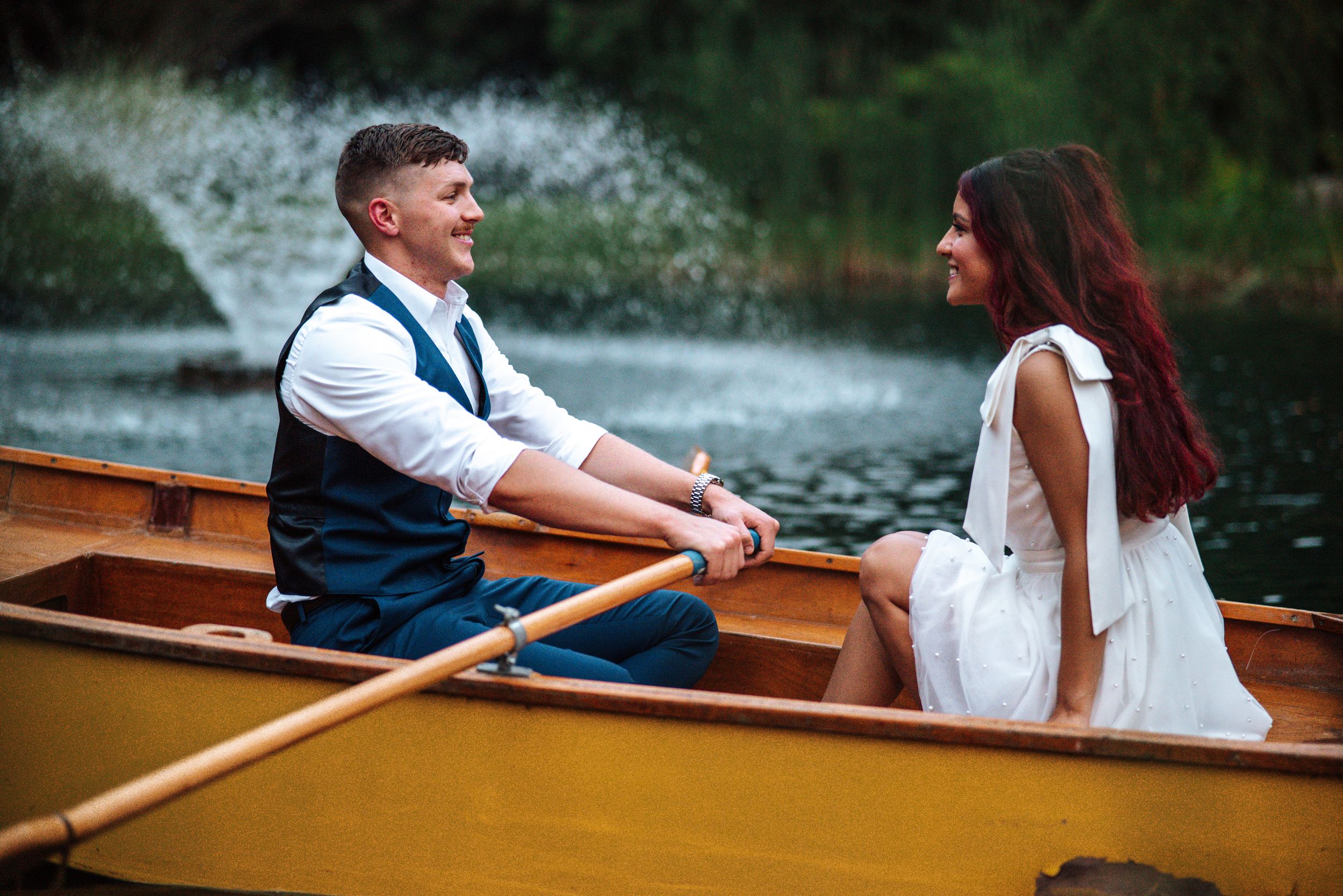 Romantic couple in formal attire sitting together in wooden rowboat on serene lake during sunset.