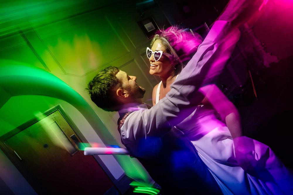 Wedding guests dancing and celebrating on a dance floor illuminated by colorful neon lights.
