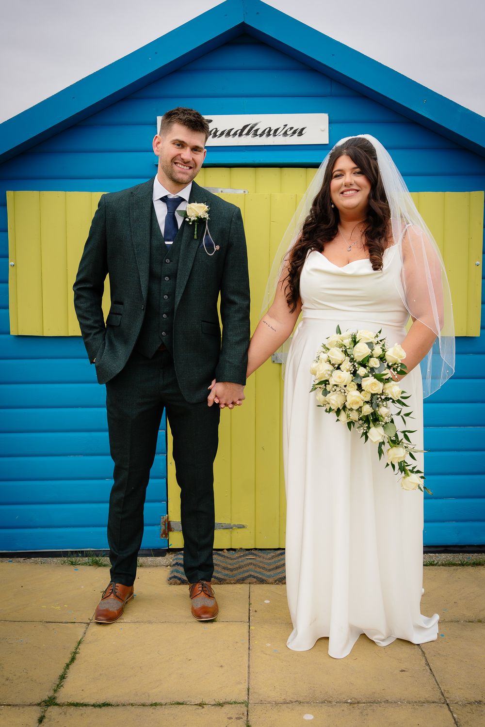 Newlyweds posing in front of bright blue and yellow beach huts in casual wedding attire.