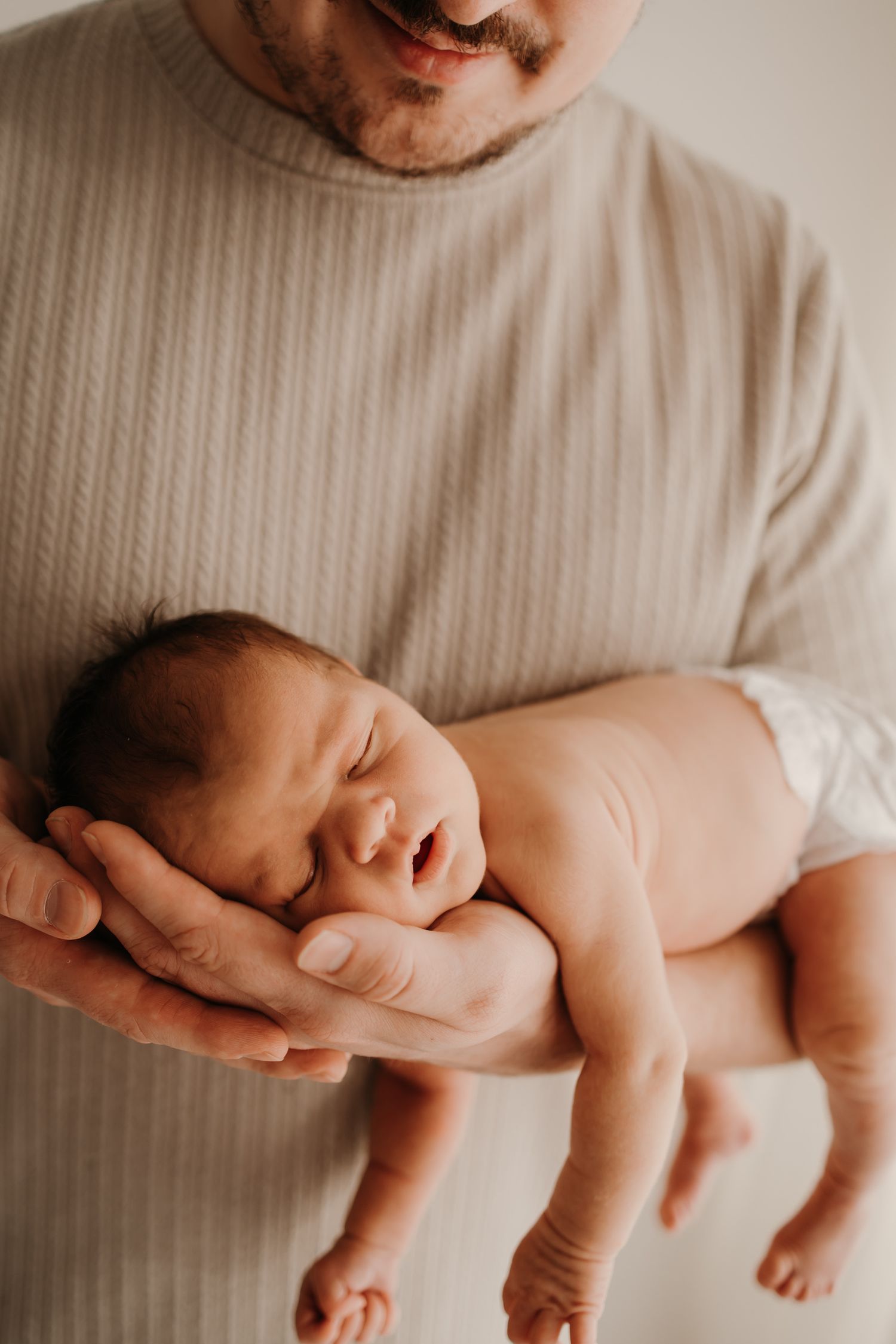 A sleeping newborn baby rests peacefully in gentle hands against a soft cream-colored background.