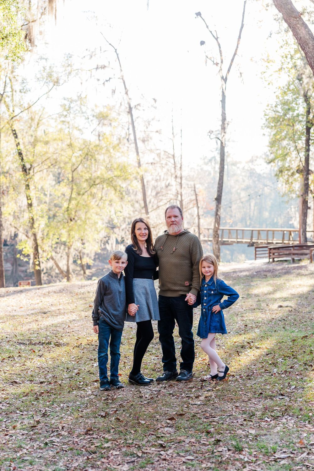 A family of four poses together on a wooded trail during autumn with fallen leaves and sunlight filtering through trees.