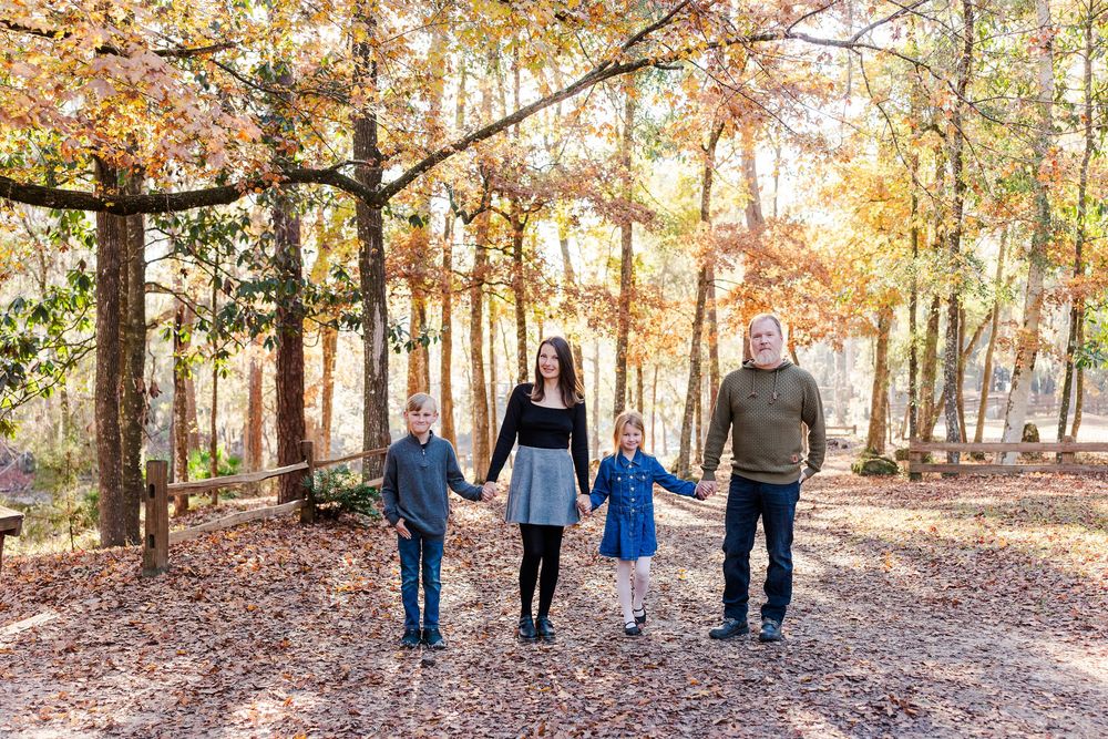 A family walks hand in hand along a sunlit forest path covered in fallen autumn leaves.