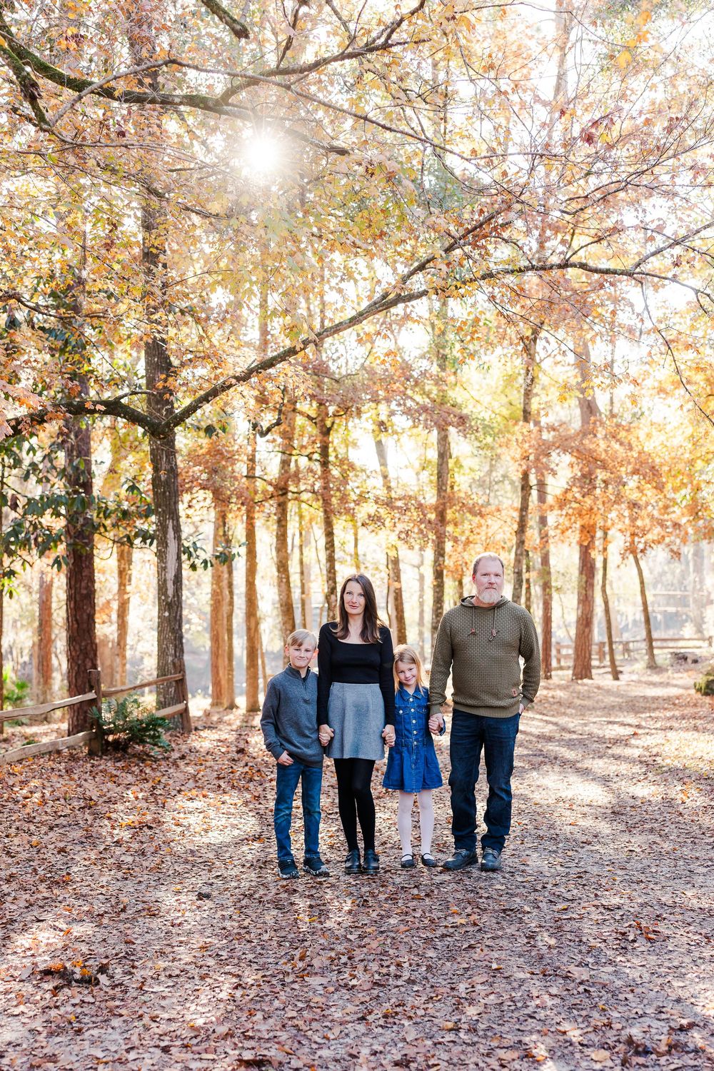 Family group standing together on fallen autumn leaves in sunlit wooded park.