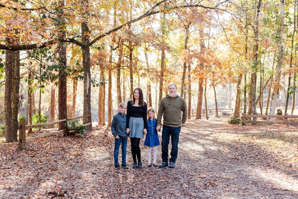 Family walking together on a sunlit autumn forest trail with golden leaves and tall trees.