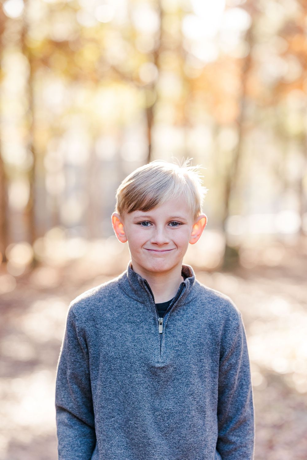 A child wearing a gray quarter-zip sweater poses outdoors in a wooded setting with soft natural lighting.