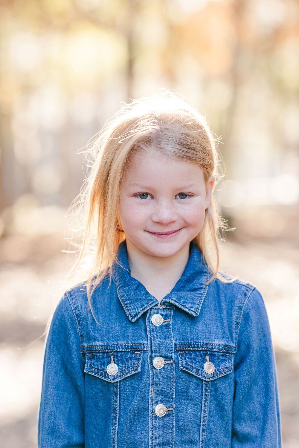 A young child wearing a denim jacket smiles at the camera with autumn foliage blurred in the background.