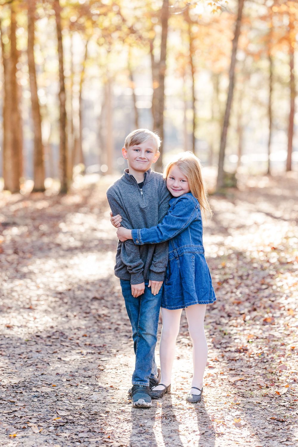 Two children in casual clothing embrace while standing on a forest path with autumn trees and golden sunlight behind them.