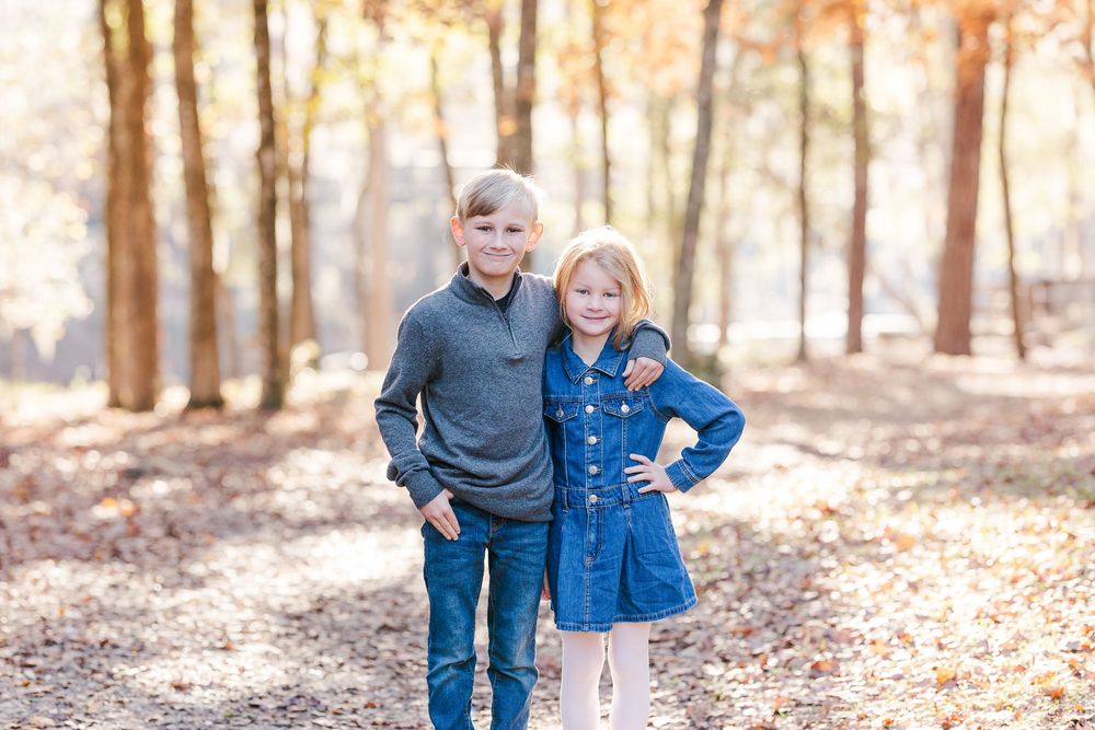 Two children stand together on a wooded path during autumn with sunlight filtering through the trees.