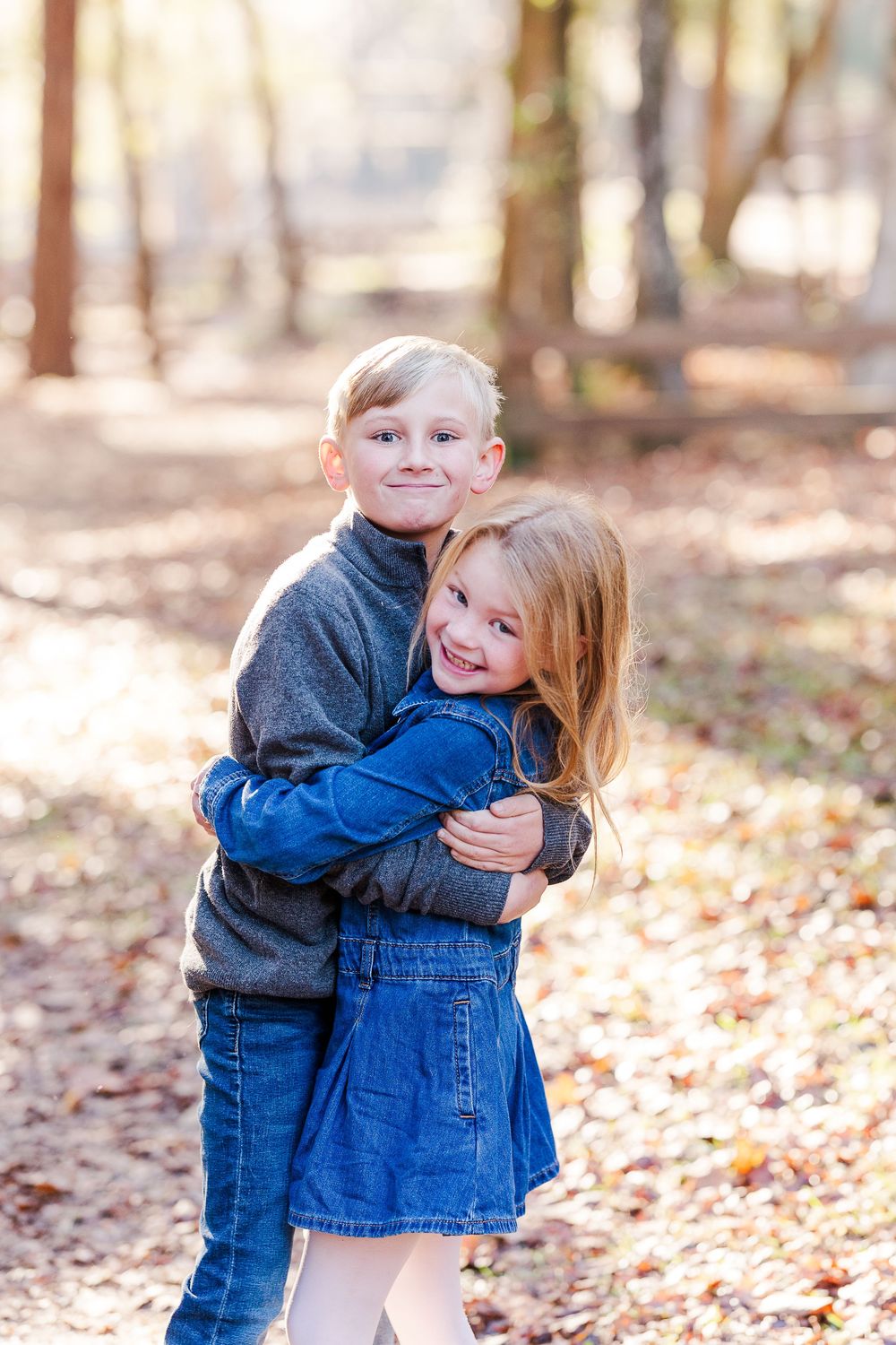 Two young children in denim and blue sweaters hug and smile together in a sunlit autumn forest.