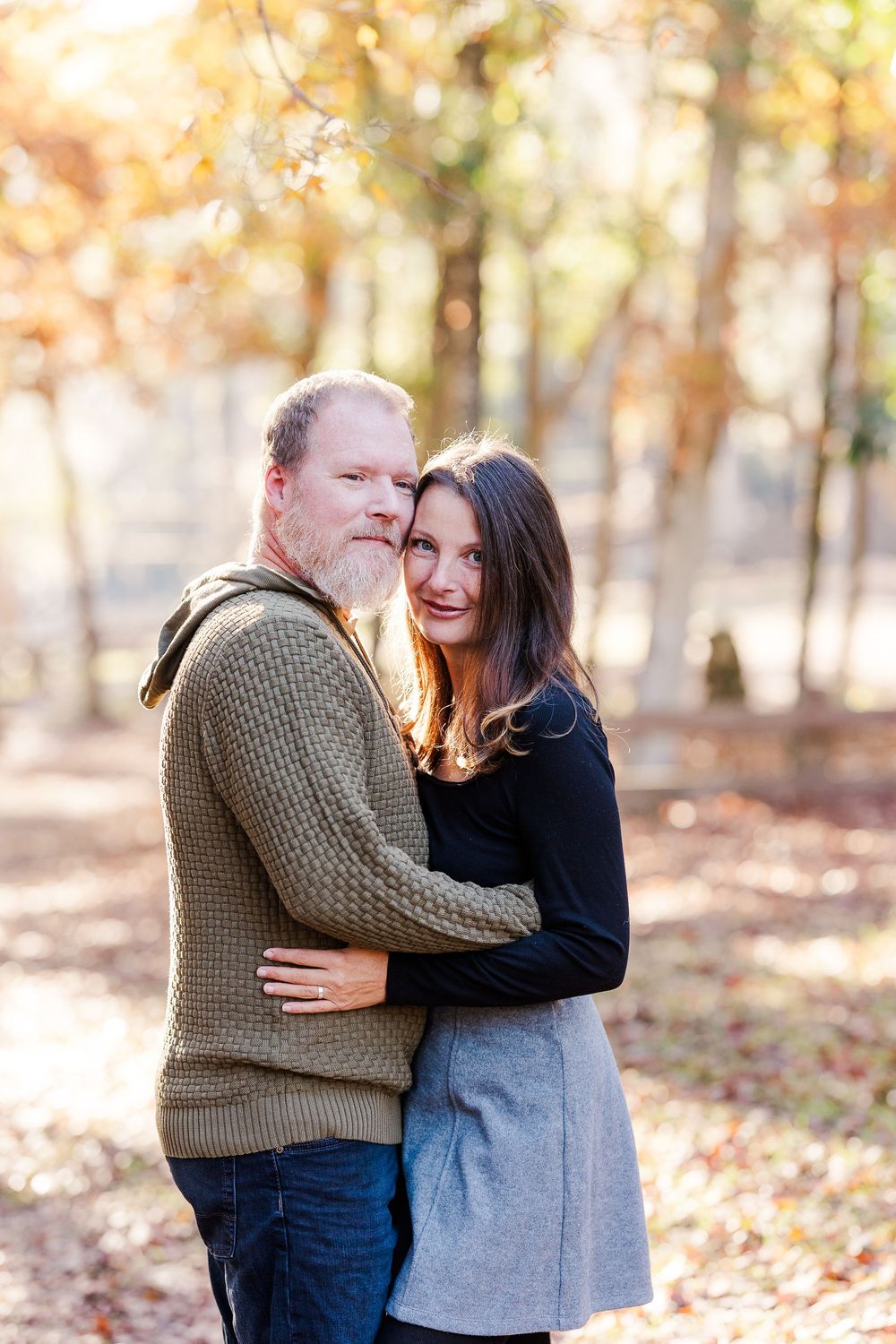 Couple laughing and embracing outdoors in autumn forest with fallen leaves on ground during photo session.