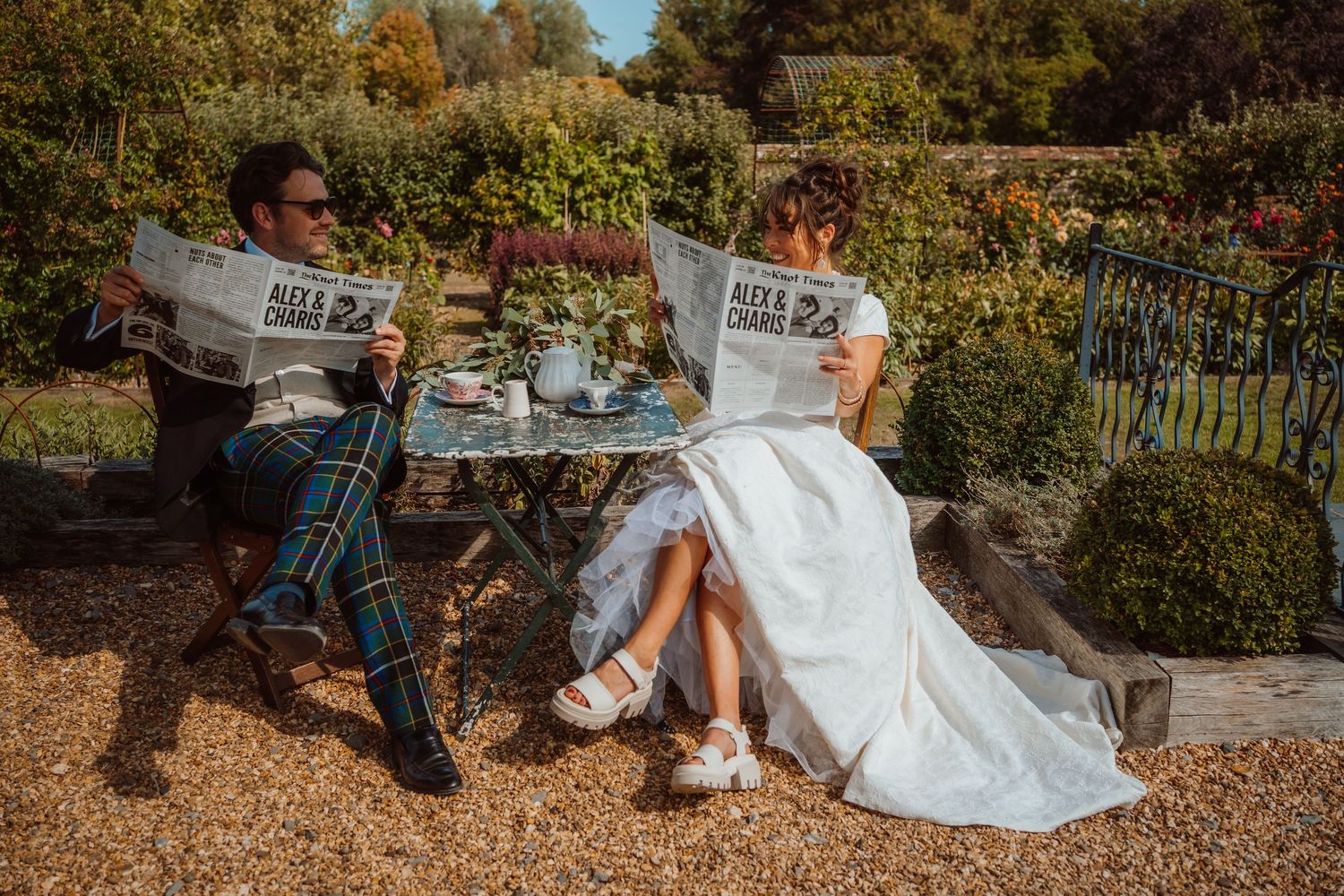 A couple in wedding attire sits on a wooden bench in a garden setting during autumn.