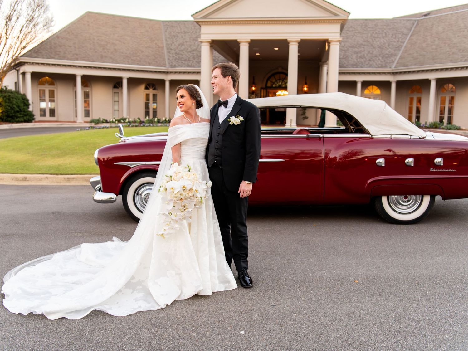 Elegant wedding couple poses in front of a classic burgundy vintage car.