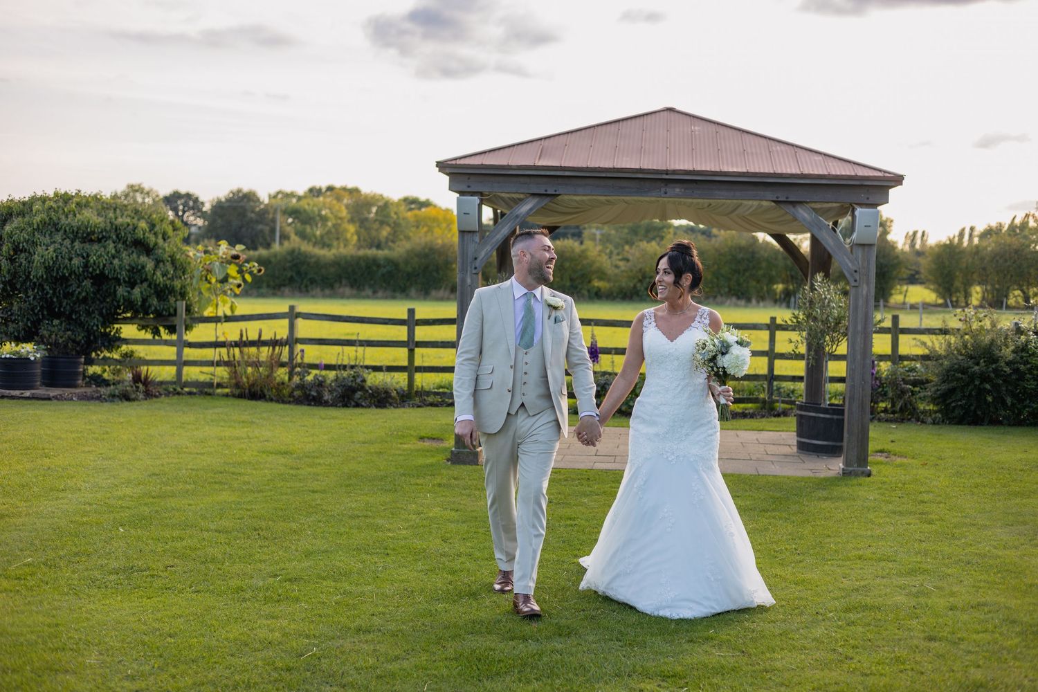 A couple in wedding attire walks across a manicured lawn with a wooden gazebo in the background at sunset.
