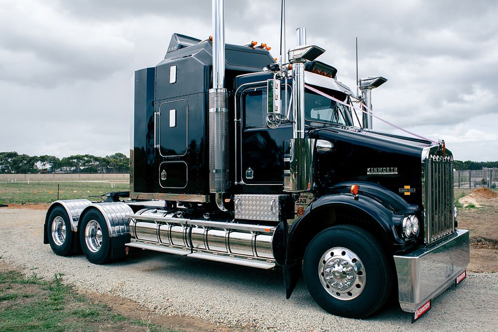 A black semi truck with custom chrome details and polished wheels sits parked on a dirt road under cloudy skies.