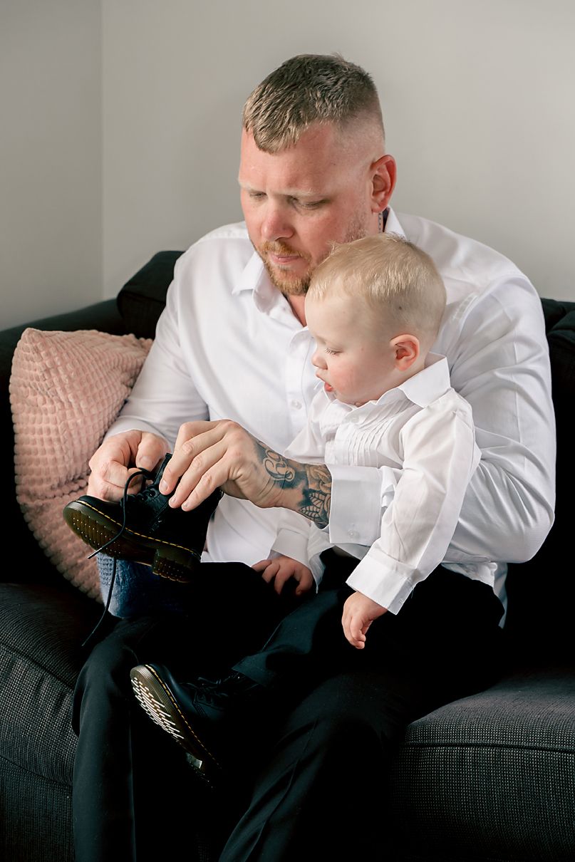A parent and young child sit on a dark couch looking at a camera or device together, both wearing white shirts.