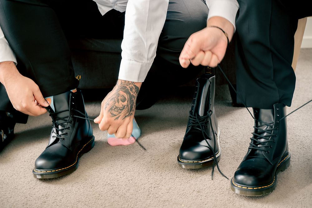 Close-up view of hands with tattoos tying black leather boots on a carpeted floor.