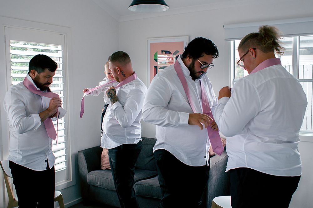 A group of people in white dress shirts and pink ties help each other get ready in a bright room.