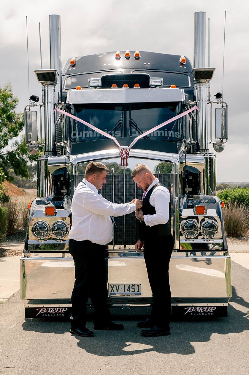 Two people in formal attire stand in front of parked semi trucks performing a celebratory high-five gesture.