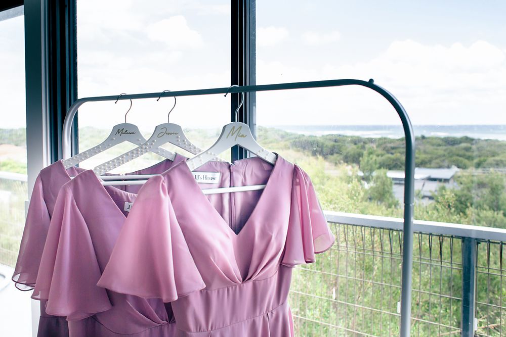 Pink bridesmaid dress hanging on clothing rack with scenic outdoor view through window in background.