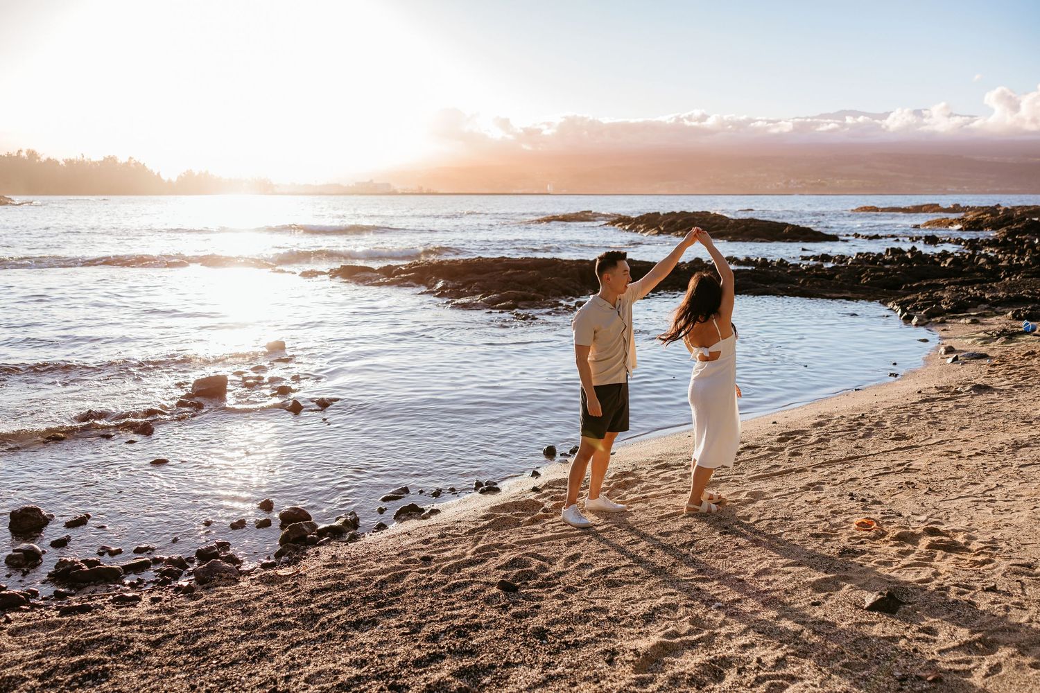 A couple walks along a rocky beach shoreline at sunset while waves crash against the coast.
