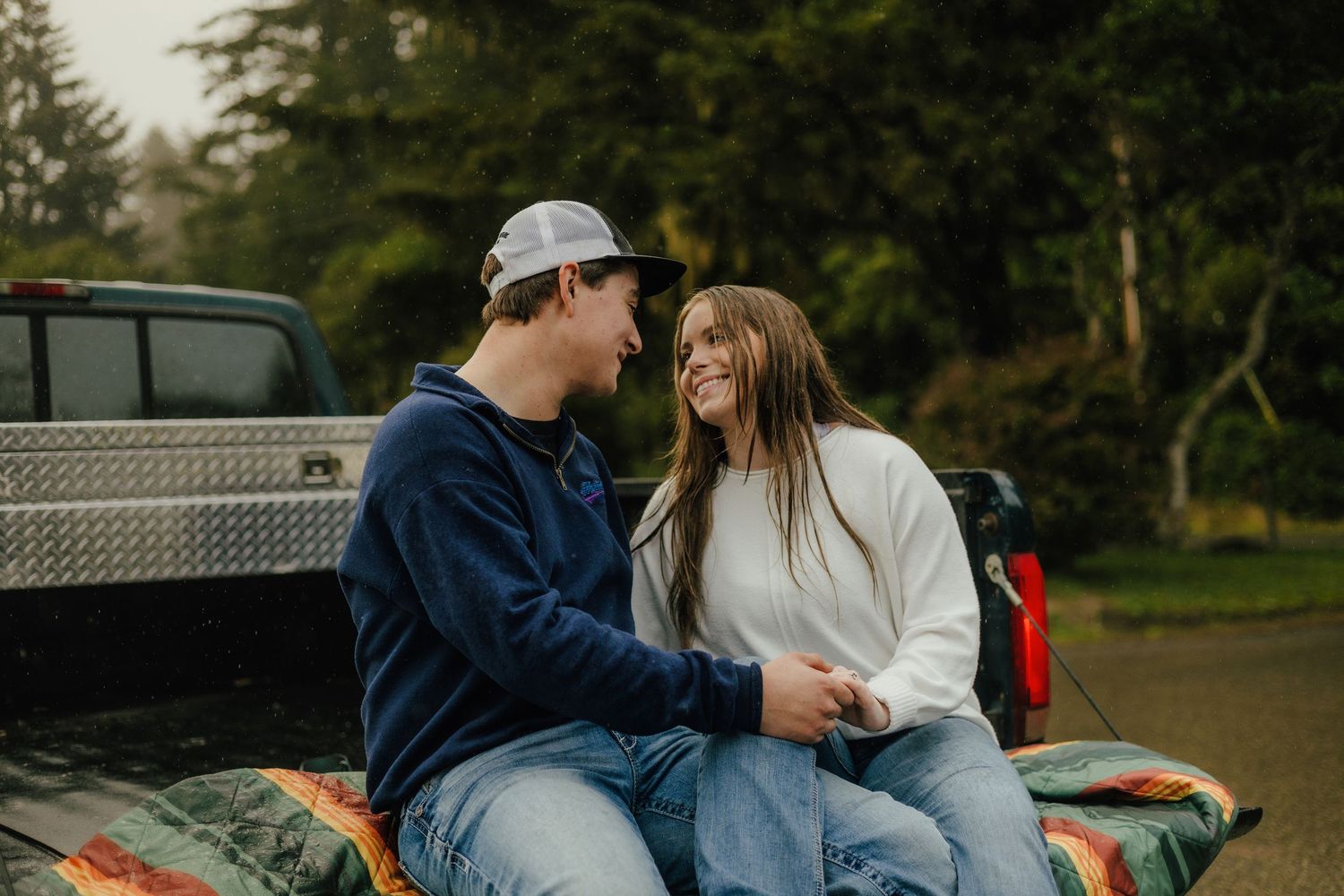 A couple shares a romantic moment while sitting in the back of a pickup truck in a wooded setting.