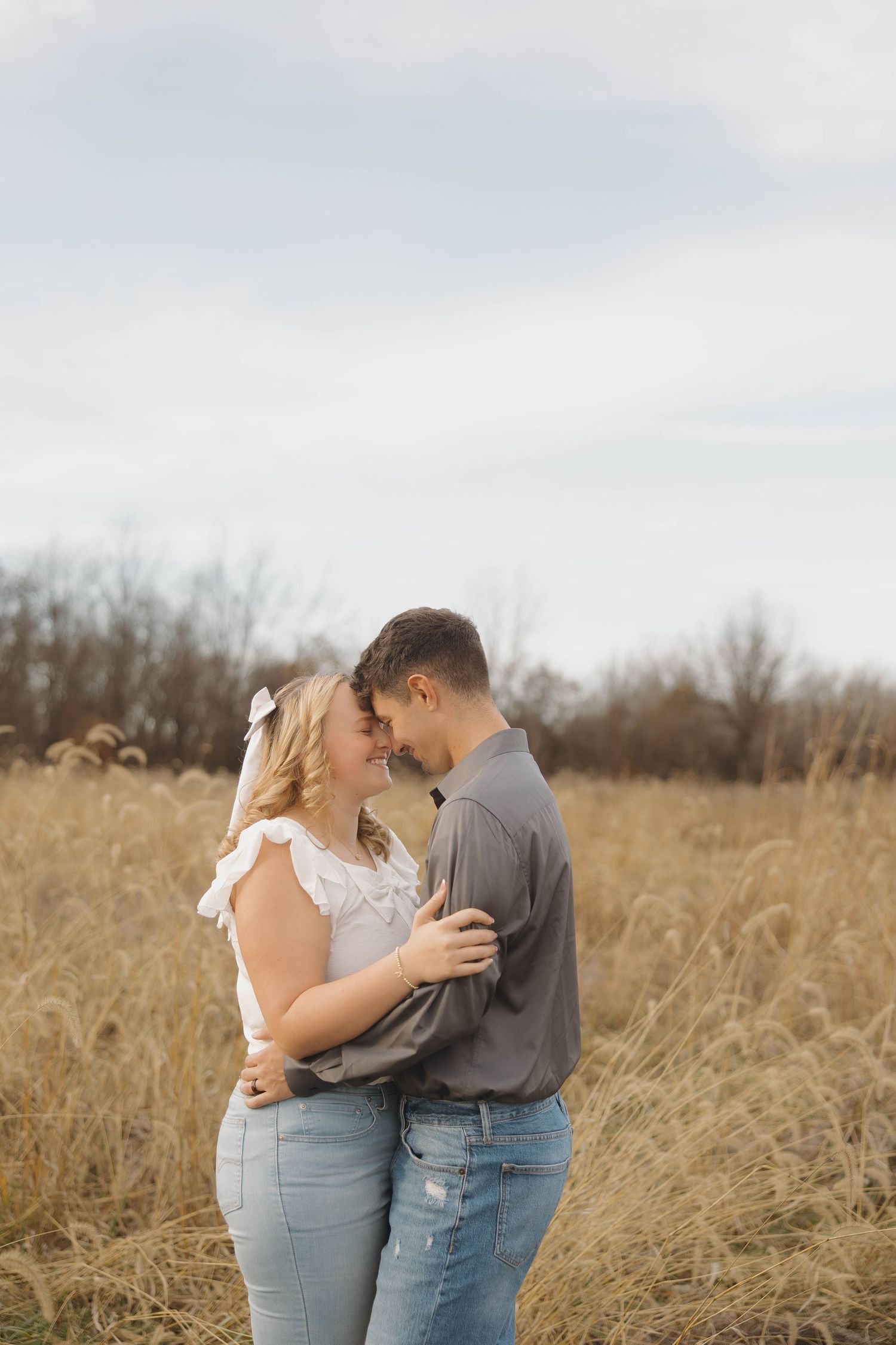 A couple embraces intimately in a golden autumn field with tall grass and bare trees in the background.