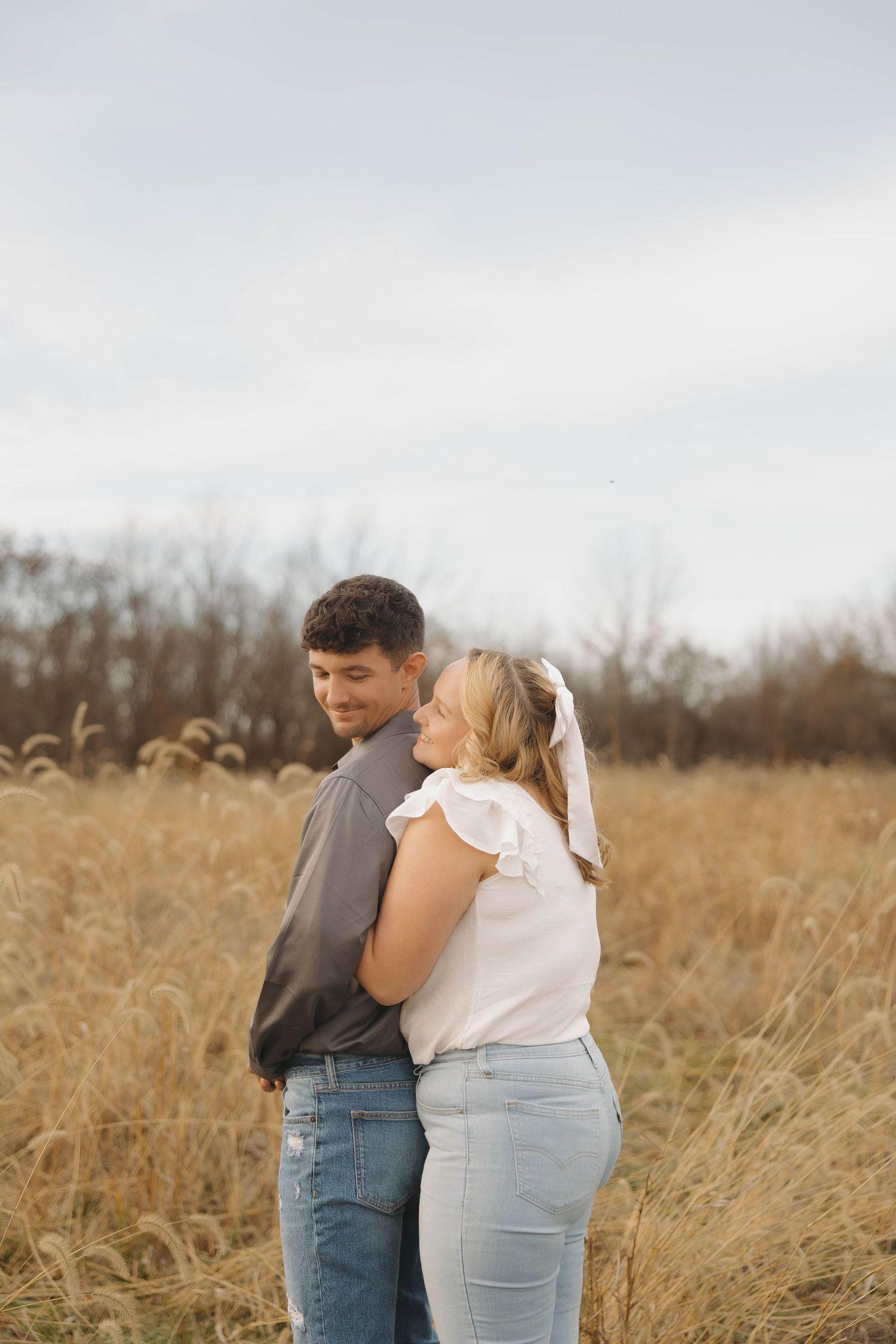 A couple embraces while standing in a golden autumn field with tall grass and trees in the background.