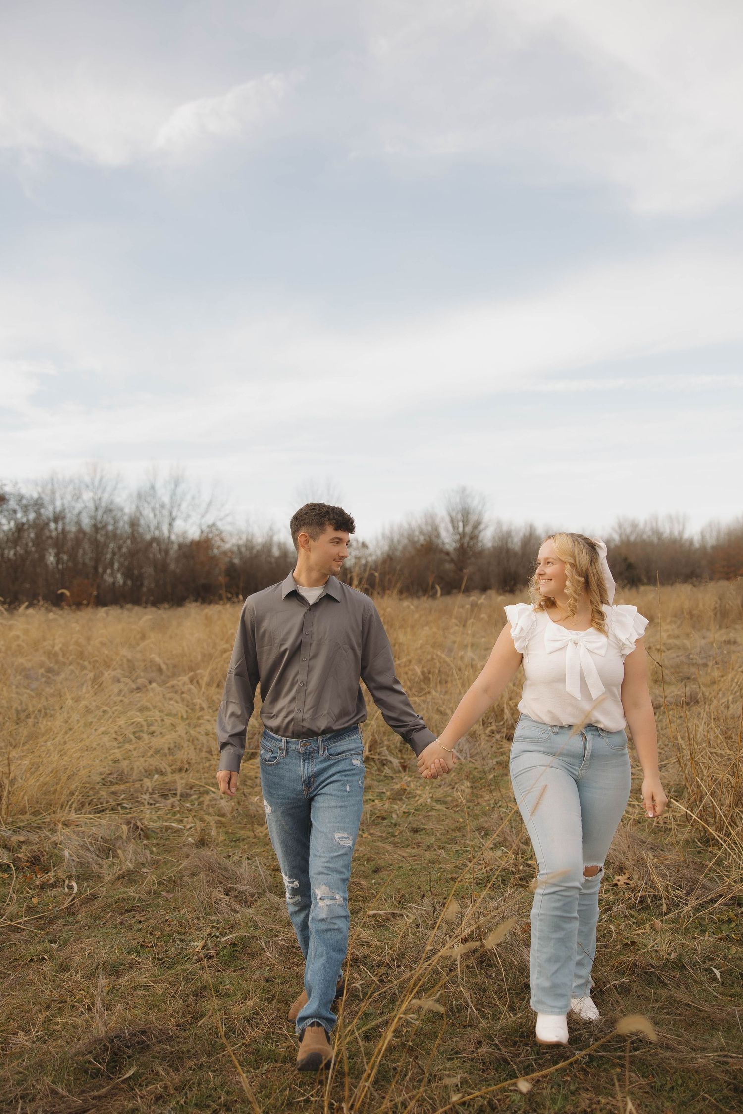 A couple walks hand in hand through a golden autumn field with dried grass and bare trees in the background.