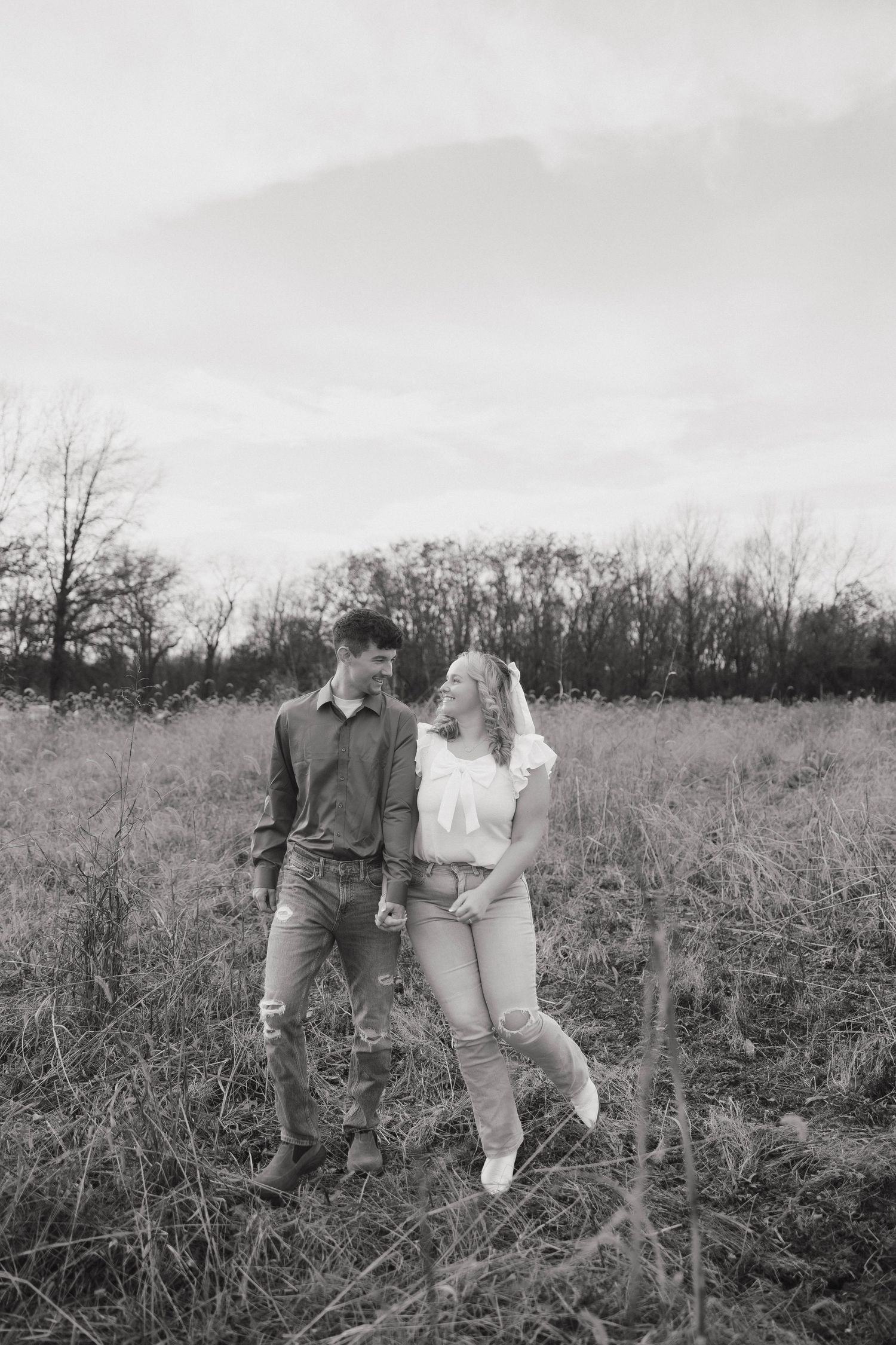 A romantic black and white photo of a couple standing together in a rustic field with trees.